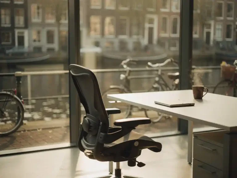Ergonomische bureaustoel bij een modern werkstation in Amsterdam, dampende koffiemok naast een notitieboek, ochtendlicht door hoge ramen met uitzicht op een gracht.